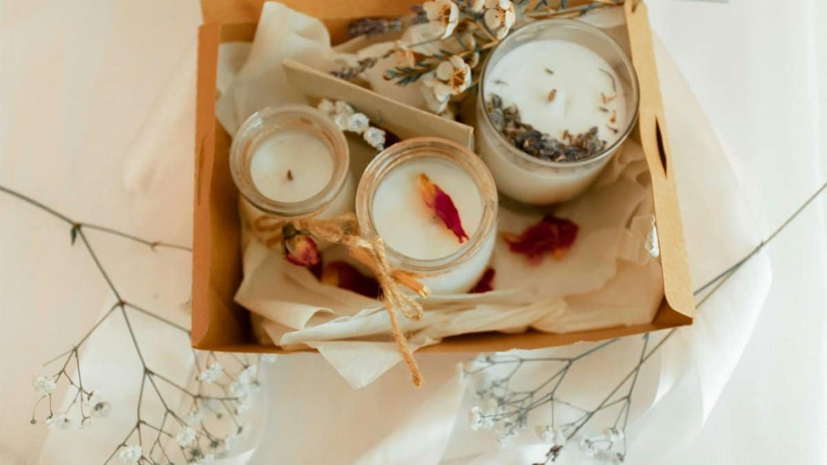 From above of cardboard present box with scented candles and envelope decorated with small flowers and twigs on white textile