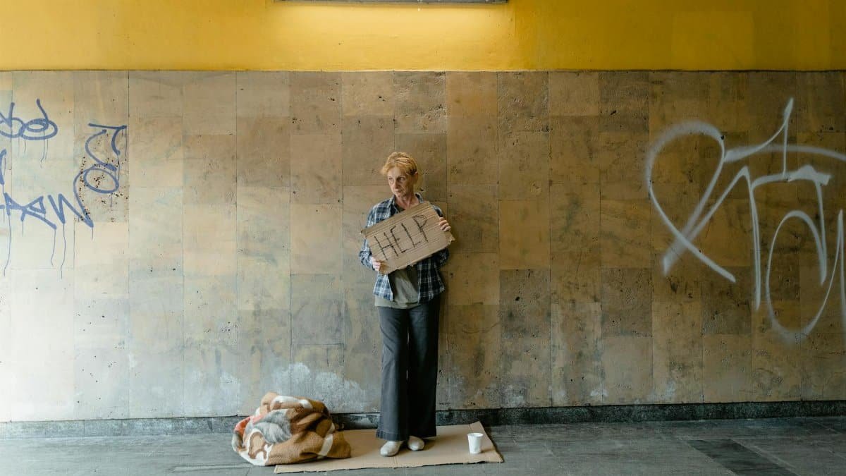 A senior woman stands with a 'HELP' sign in an urban area, symbolizing poverty and homelessness.