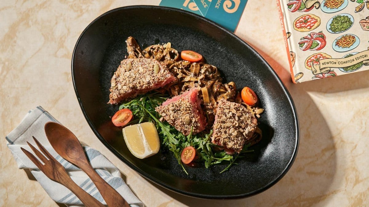 A nut-crusted tuna steak dish with cherry tomatoes and lemon served on a black plate.