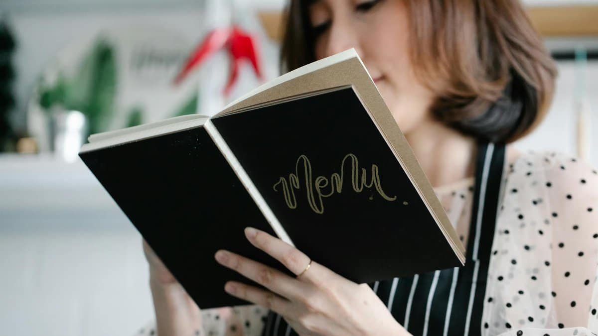 A chef in a polka dot blouse holds a menu book in a stylish restaurant, ready to present the dishes.