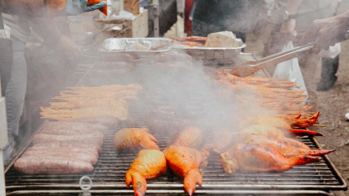 Street market scene with various meats grilling on a barbecue under smoky conditions.
