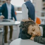 A sad child rests on a classroom desk while peers talk, symbolizing loneliness and school exclusion.