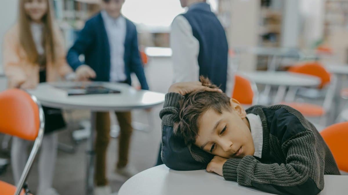 A sad child rests on a classroom desk while peers talk, symbolizing loneliness and school exclusion.