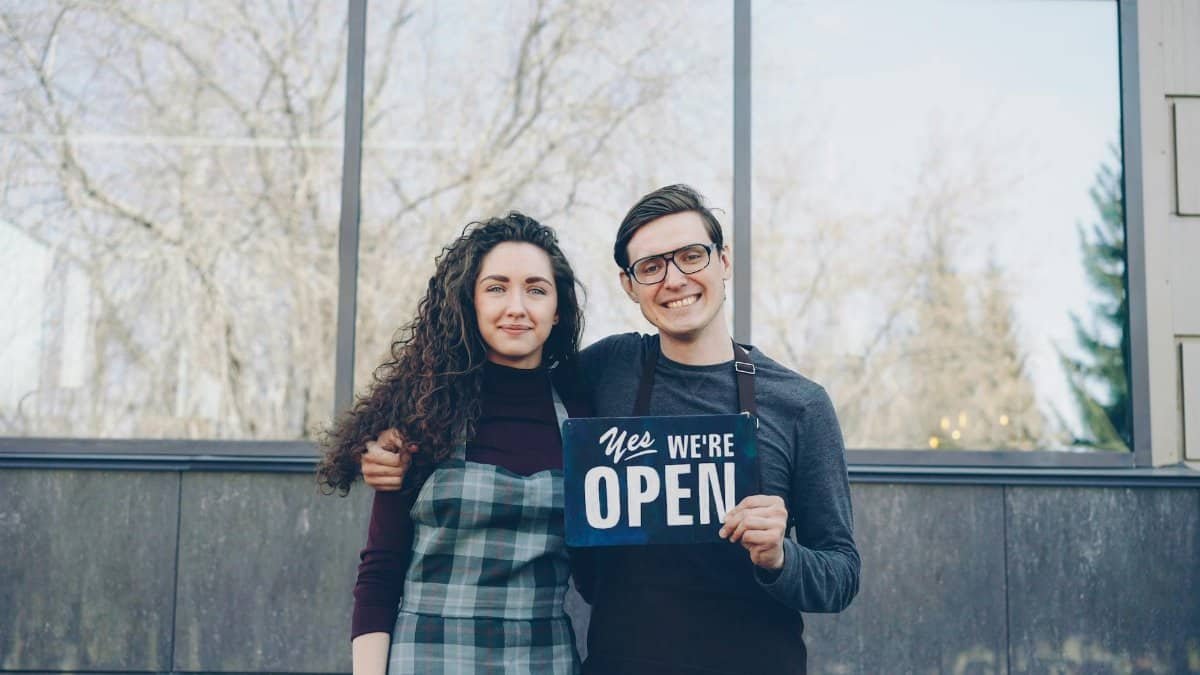 Smiling business owners standing outside with an open sign, ready to welcome customers.