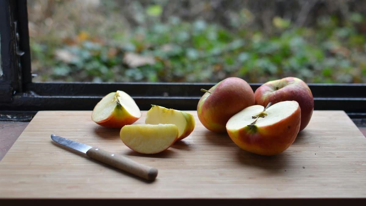 High angle of cut red apples placed on wooden cutting board with knife near window