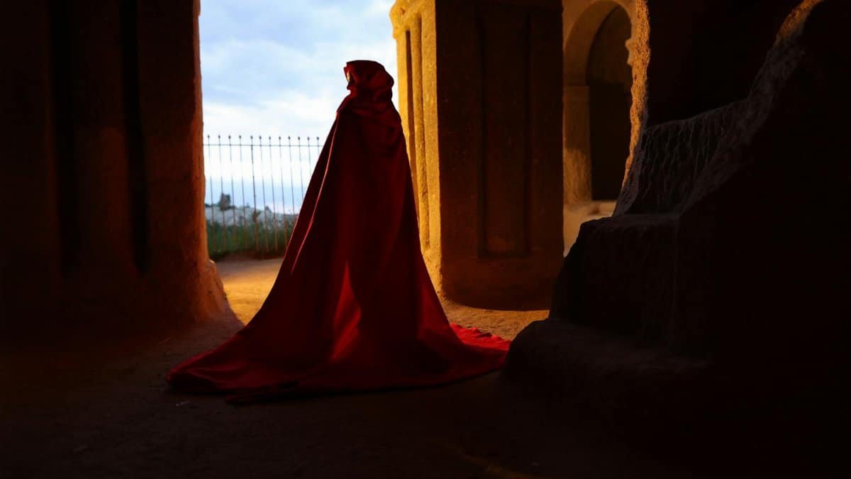 A cloaked figure in red silhouetted against the light from an ancient stone structure.