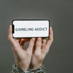 Chained hands hold a phone displaying the text 'Gambling Addict', highlighting addiction.