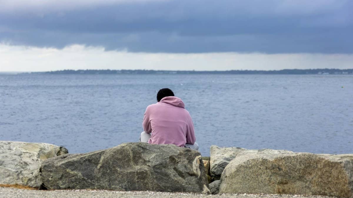 Person in hoodie sitting on rocks by the ocean shore with cloudy sky.