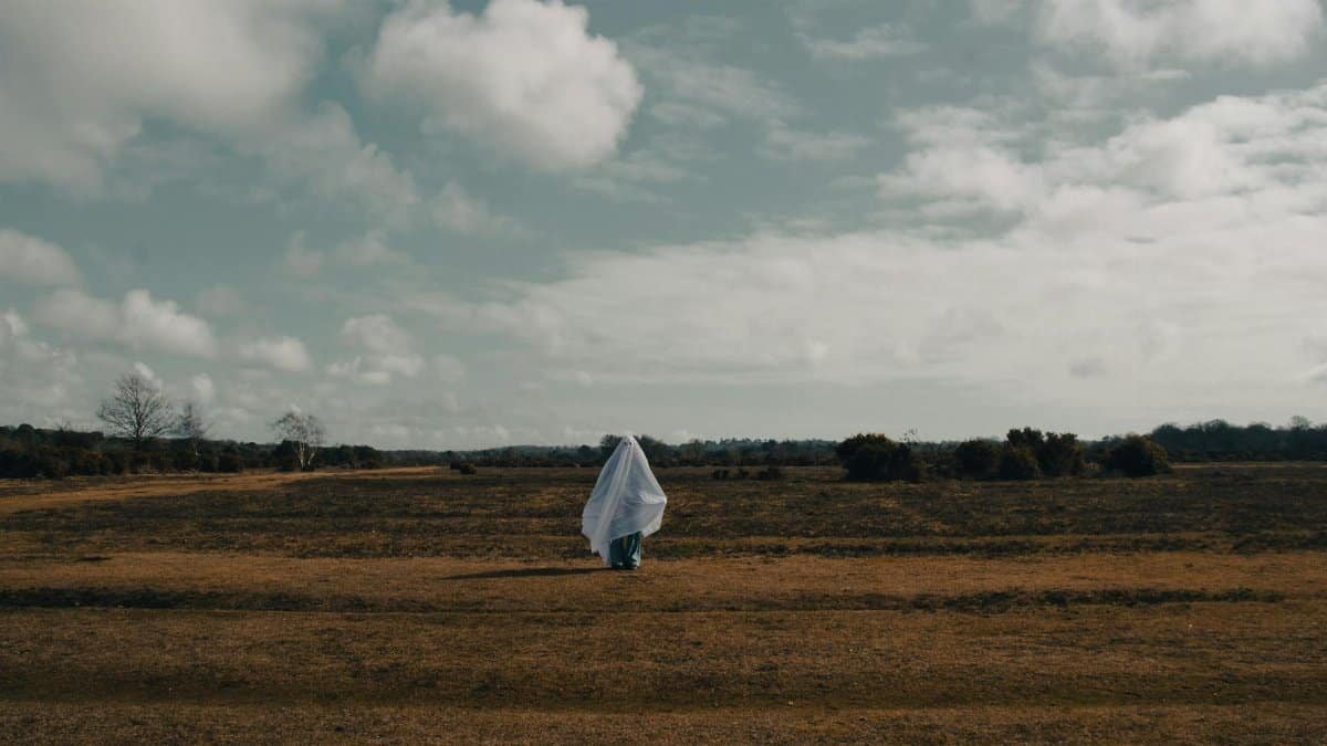 A solitary figure in a ghostly costume stands in a rural field in England under a cloudy sky.