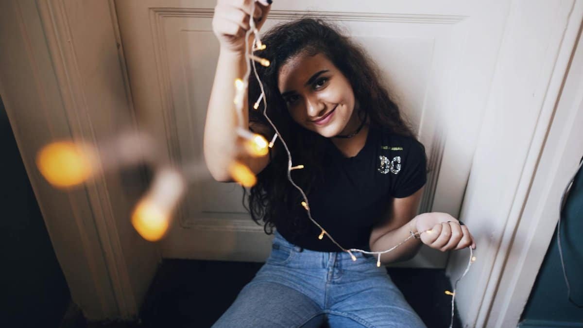 High angle of optimistic young female in casual clothes holding light garland and looking at camera while decorating room for party