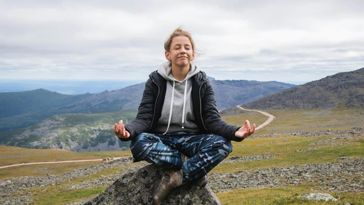 Caucasian woman meditating outdoors on a rock in a serene mountainous area.