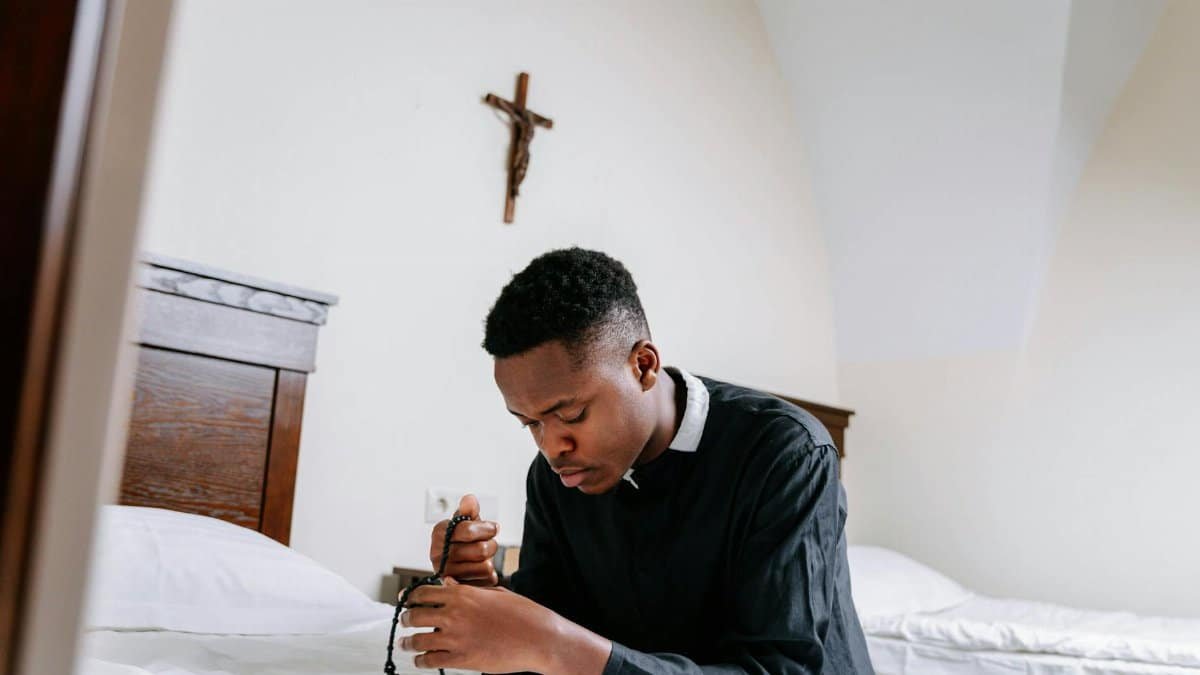 A young man kneeling in prayer holding a rosary in a tranquil bedroom setting.