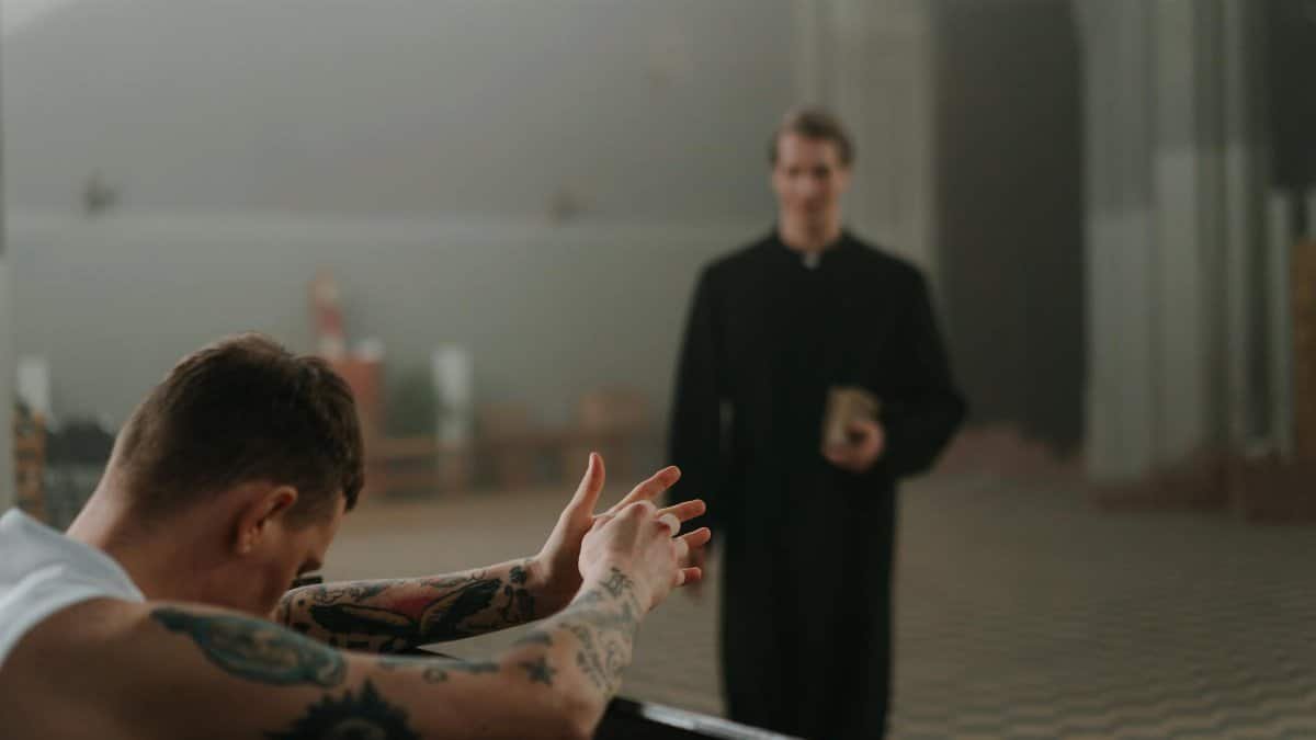 A tattooed man prays in a church as a priest observes, capturing faith and reflection indoors.