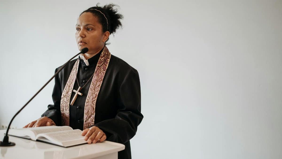 A female pastor delivers a sermon at a church podium with a Bible and microphone.