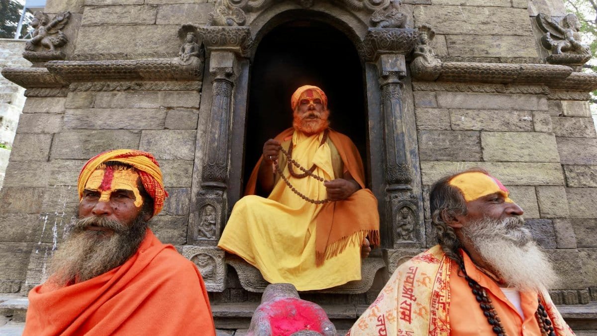 Three sadhus in vivid orange and yellow garb at a historic stone temple, showcasing spiritual tradition.