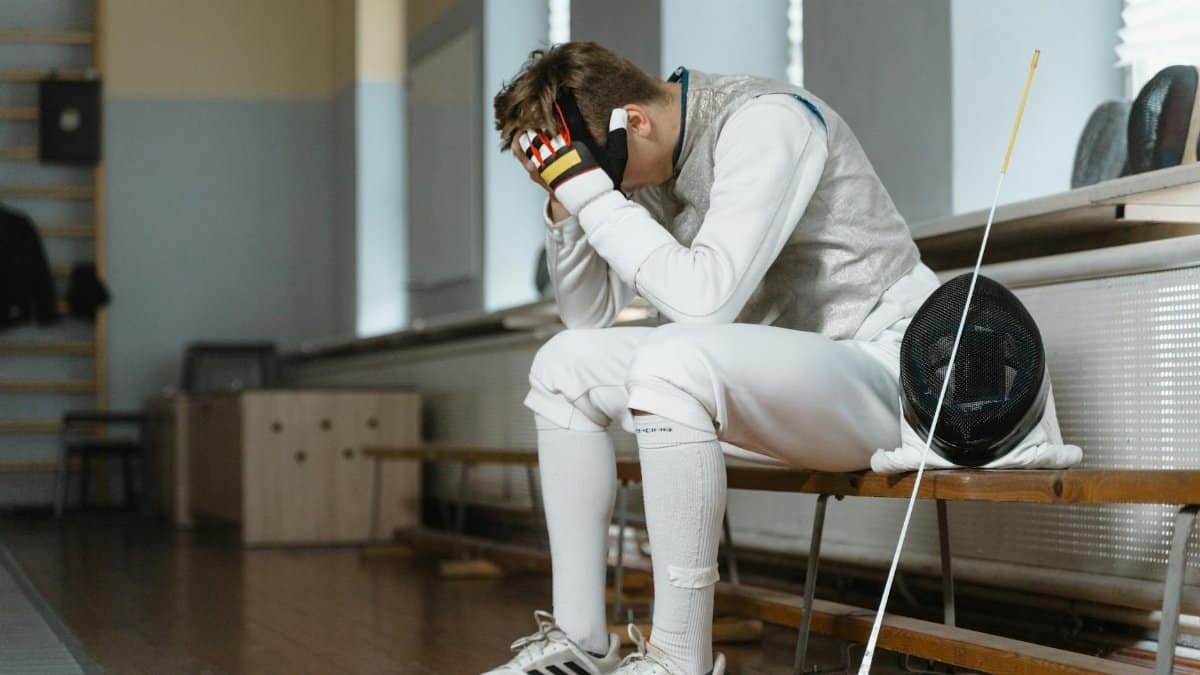 Young fencer with head in hands, showing frustration during indoor training session.