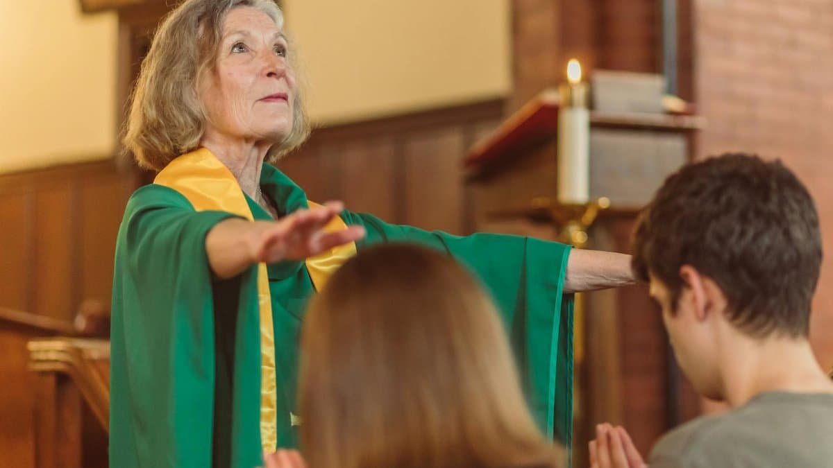 An elderly woman pastor in a green robe leading a prayer service indoors.