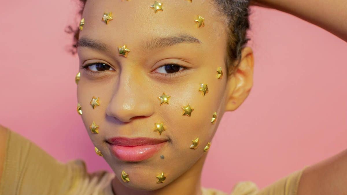 Portrait of a smiling woman with gold star stickers on her face against a pink background.
