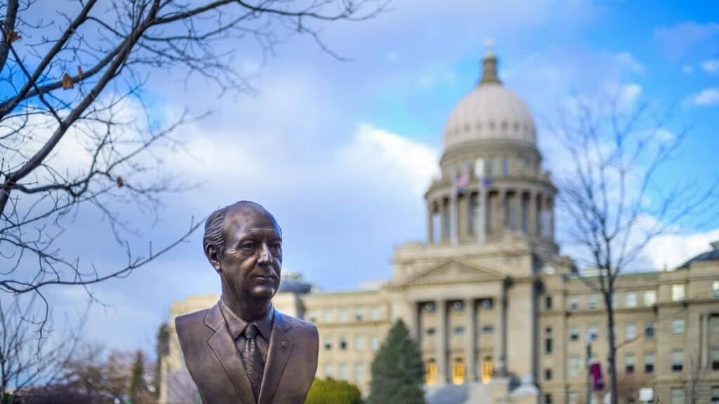 Bronze bust located in park near historic Idaho State Capitol with columns and dome against cloudy blue sky in Boise