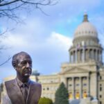 Bronze bust located in park near historic Idaho State Capitol with columns and dome against cloudy blue sky in Boise