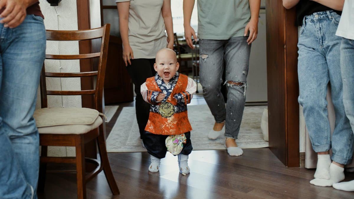 A joyful baby in traditional attire takes first steps with family support in a cozy indoor setting.
