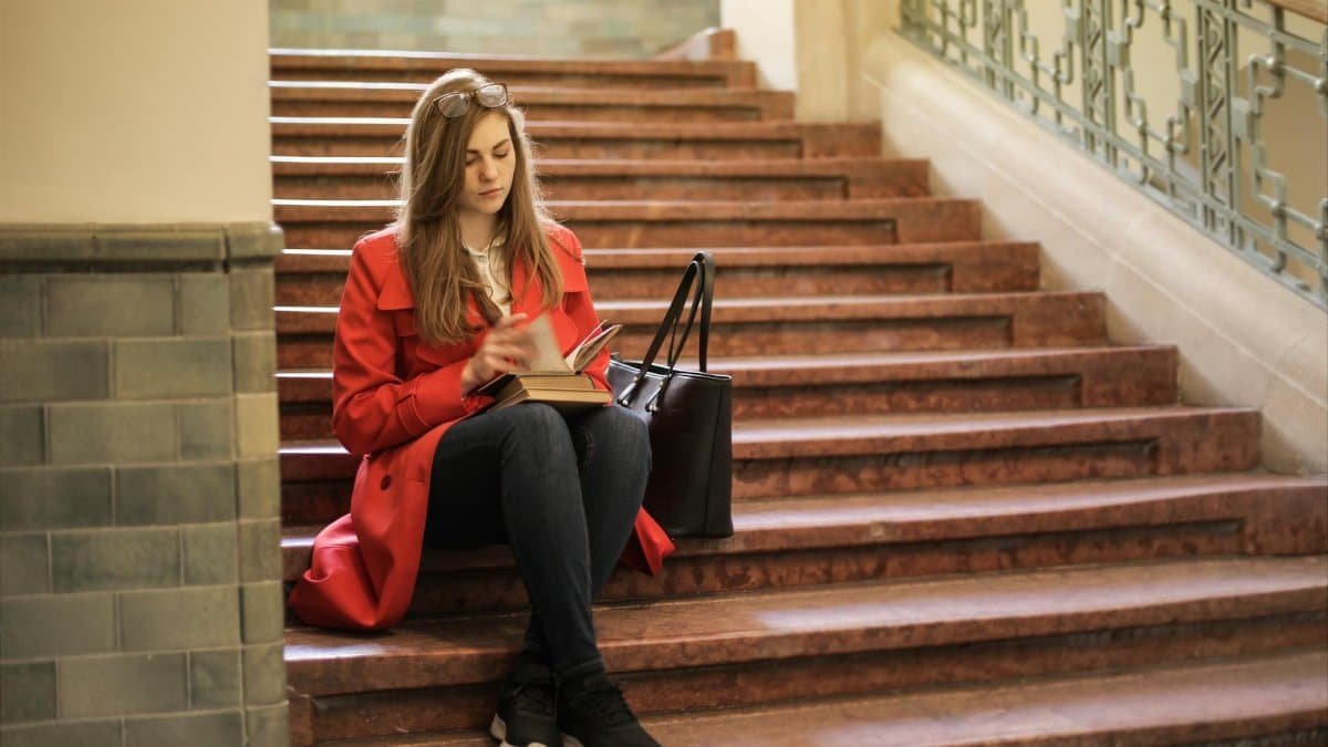 Woman in red coat reading on stairs indoors, conveying relaxation.