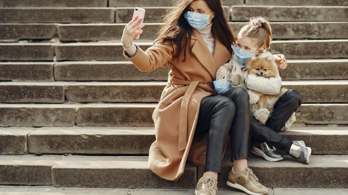 Full body of female and adorable little girl in casual clothes and medical masks sitting on steps with toy in mask and cute dog taking photo on smartphone
