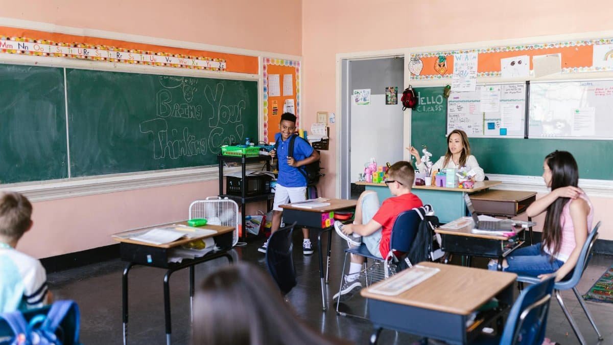 Children in a classroom with a teacher on the first day of school, chalkboard with "Bring Your Thinking Cap" message.