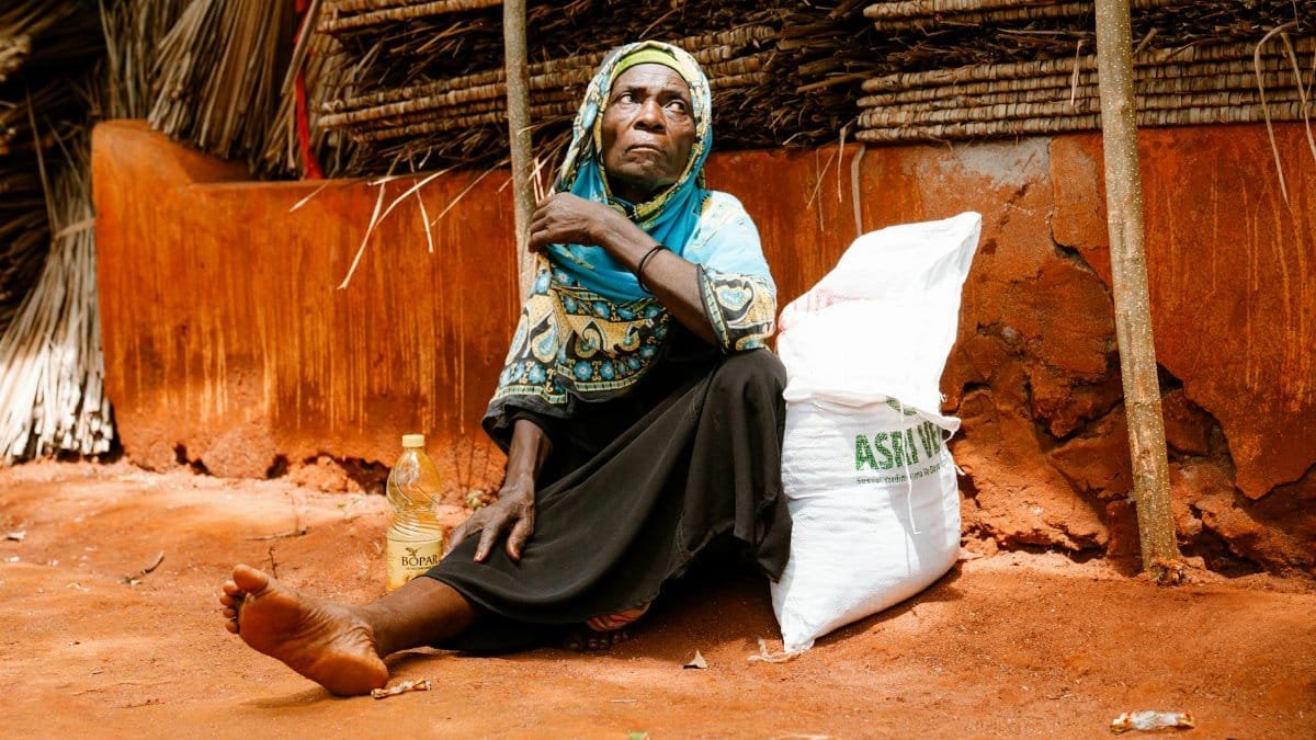An elderly woman in traditional clothing sits on the ground in a rural setting.
