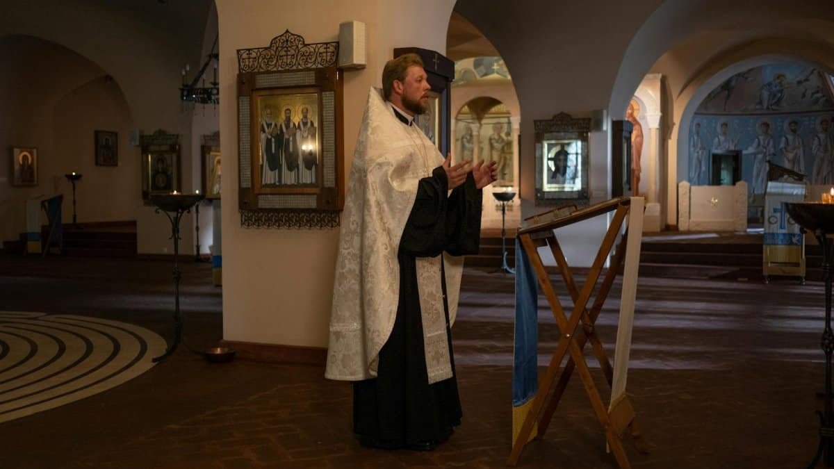 Orthodox priest in vestments standing and praying in an ornate church interior.