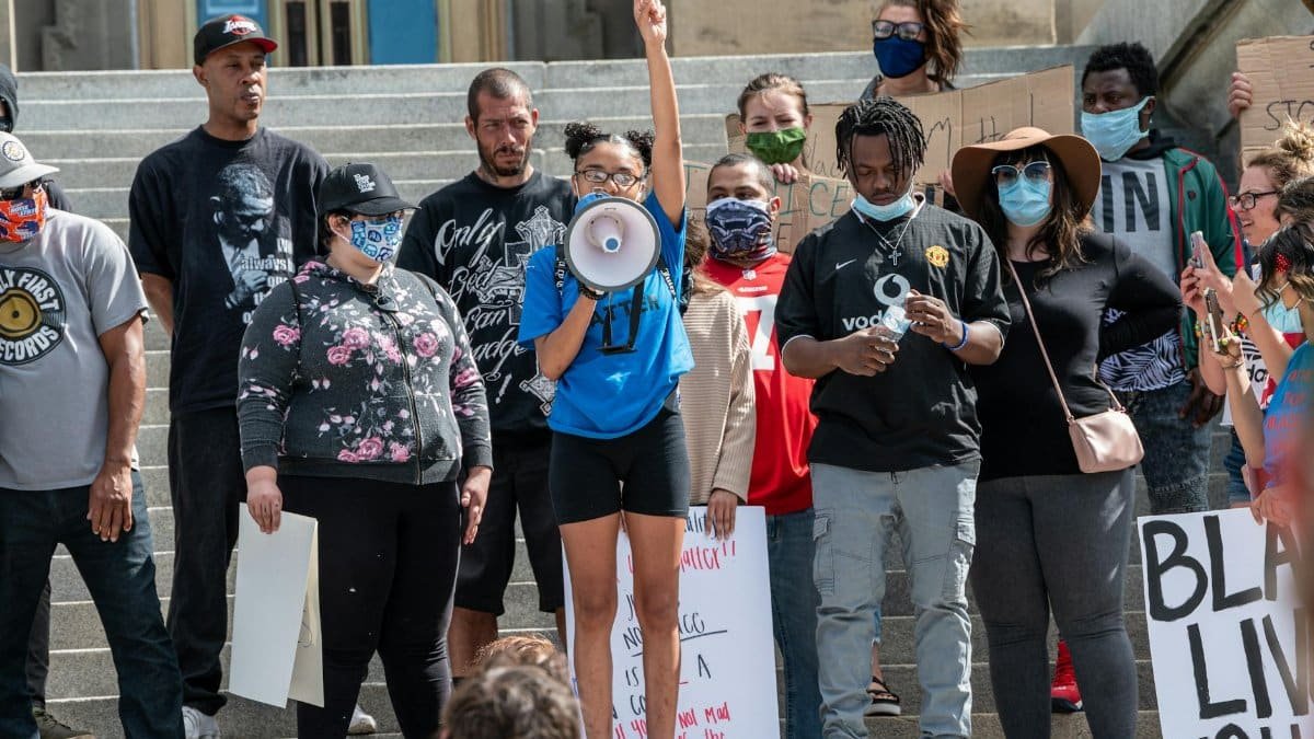 Group of young multiracial people in casual clothes and protective masks with loudspeaker and banners standing on stairs and protesting during coronavirus