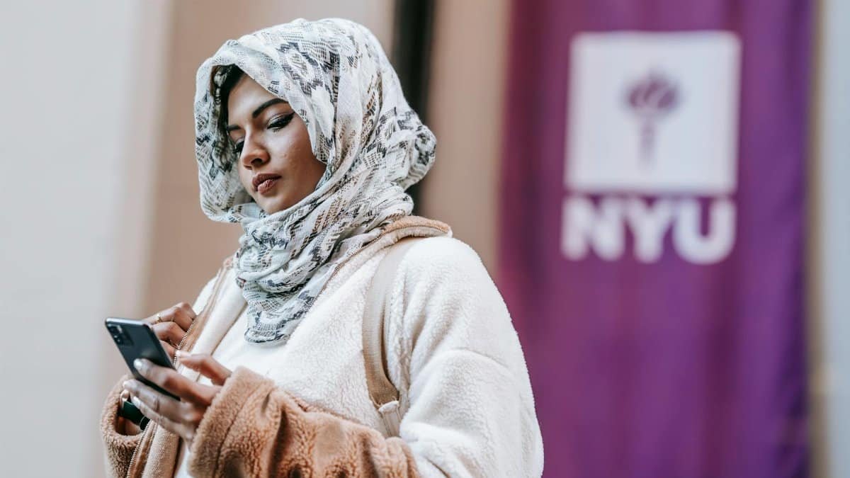 A young woman wearing a hijab stands in front of NYU, using her smartphone outdoors.