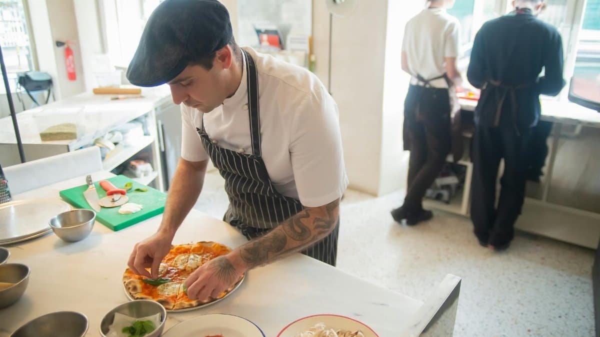 Chef preparing pizza in a professional kitchen setting, adding fresh ingredients to the dish.