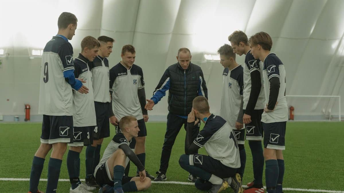 A soccer coach gives tactical advice to a youth team indoors on artificial turf.