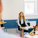 A young girl stands smiling in a bright classroom, engaged in school activities.