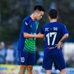 Two soccer players in blue jerseys strategize during a match in Hà Nội, Vietnam.