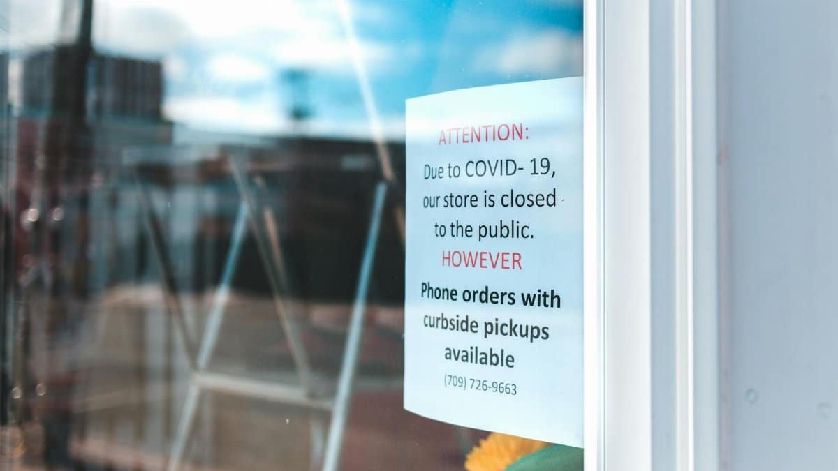 Paper sheet with warning information on glass wall of local city shop with portable ladder inside reflecting blue cloudy sky during quarantine period