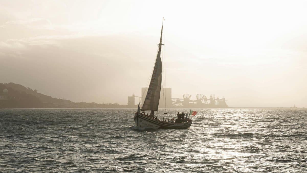 A sailboat glides on a shimmering Lisbon sea with a hazy skyline in the background.