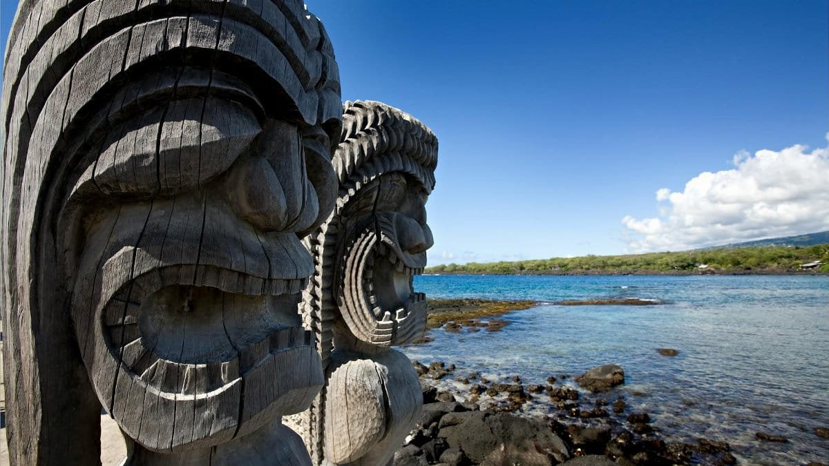 Wooden tiki statues by the ocean in Honaunau-Napoopoo, Hawaii. Bright day with rocky coastline.