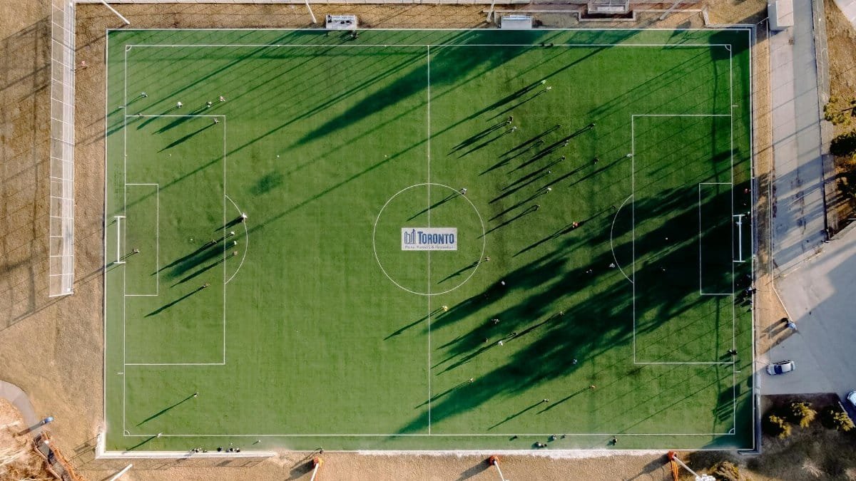 Aerial view of soccer field with anonymous sportspeople and shades against roadway on sunny day
