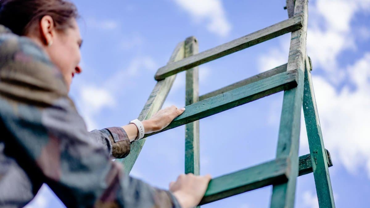 A woman climbs a green ladder under a bright blue sky. Outdoor shot.