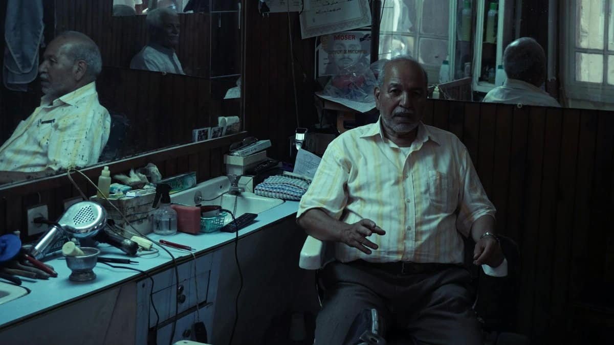 An elderly man sits in a classic barber shop surrounded by vintage grooming tools.