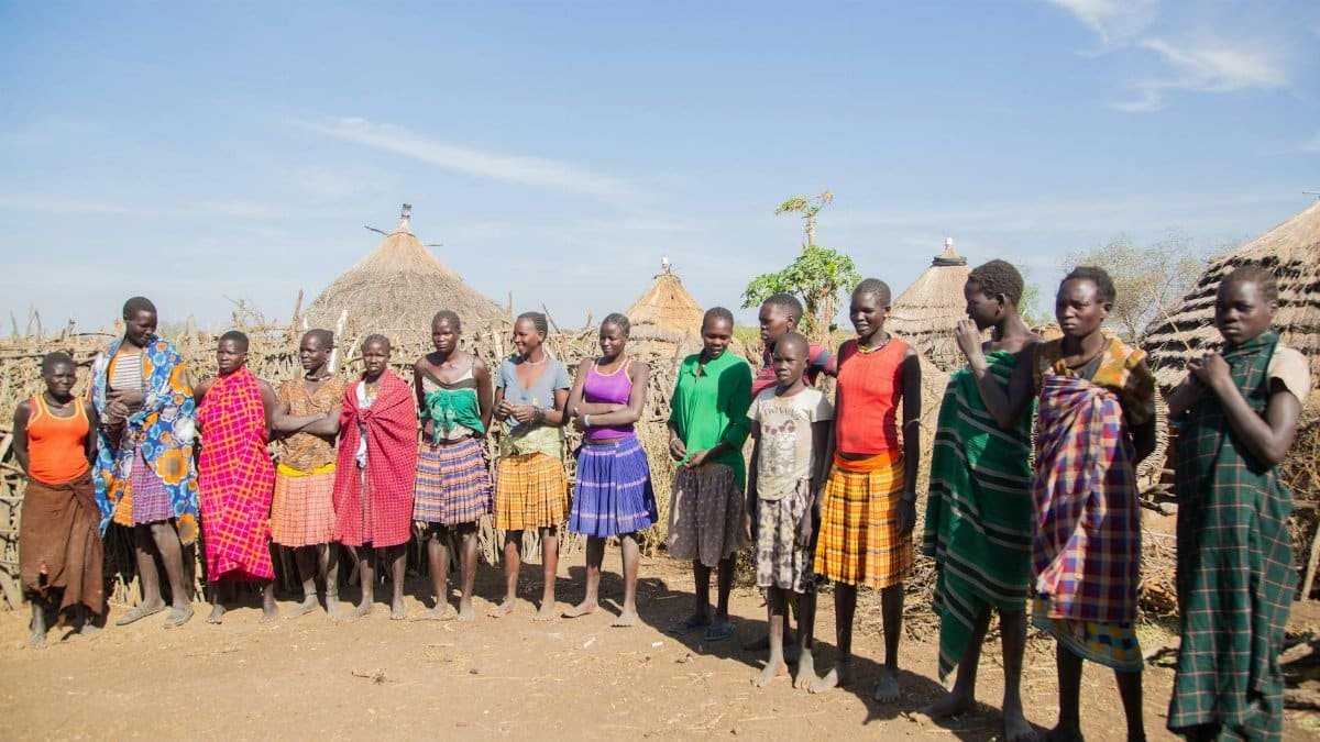 A group of people in vibrant attire gather in a rural African village under the sunny sky.