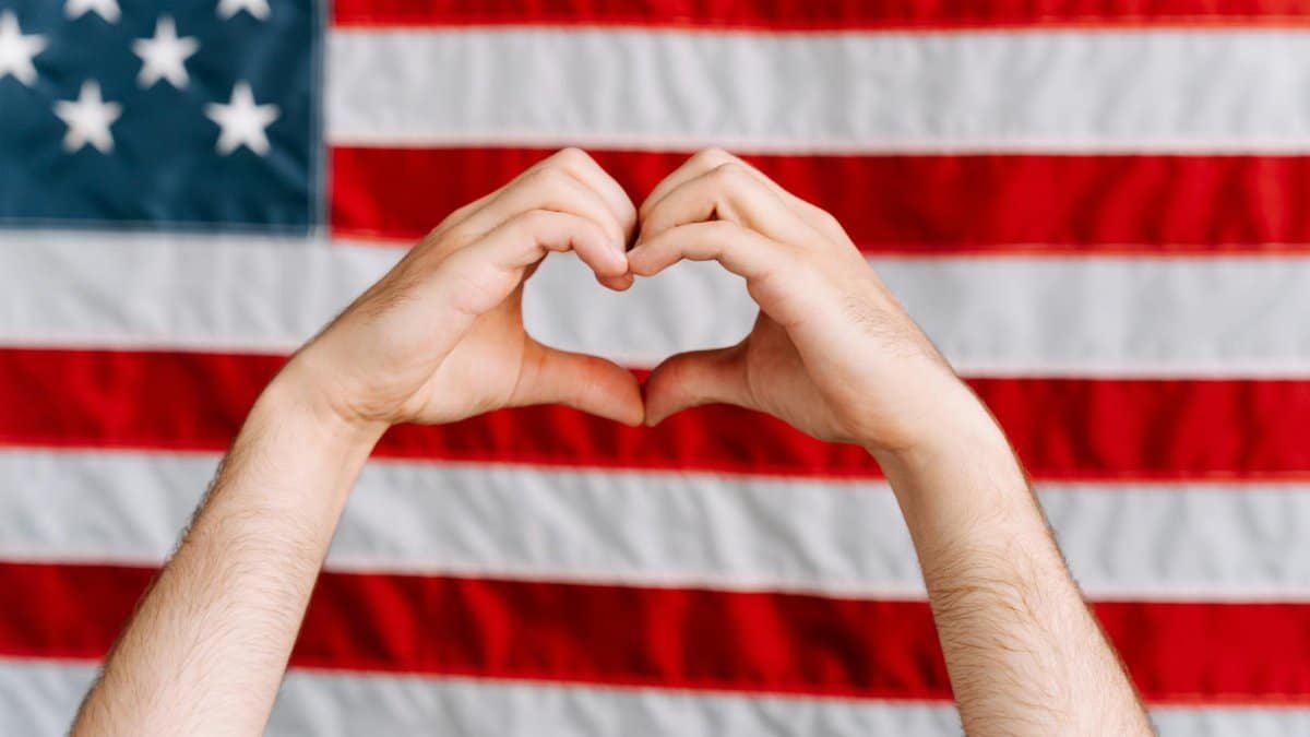 Hands forming a heart shape in front of the American flag, symbolizing love and patriotism.