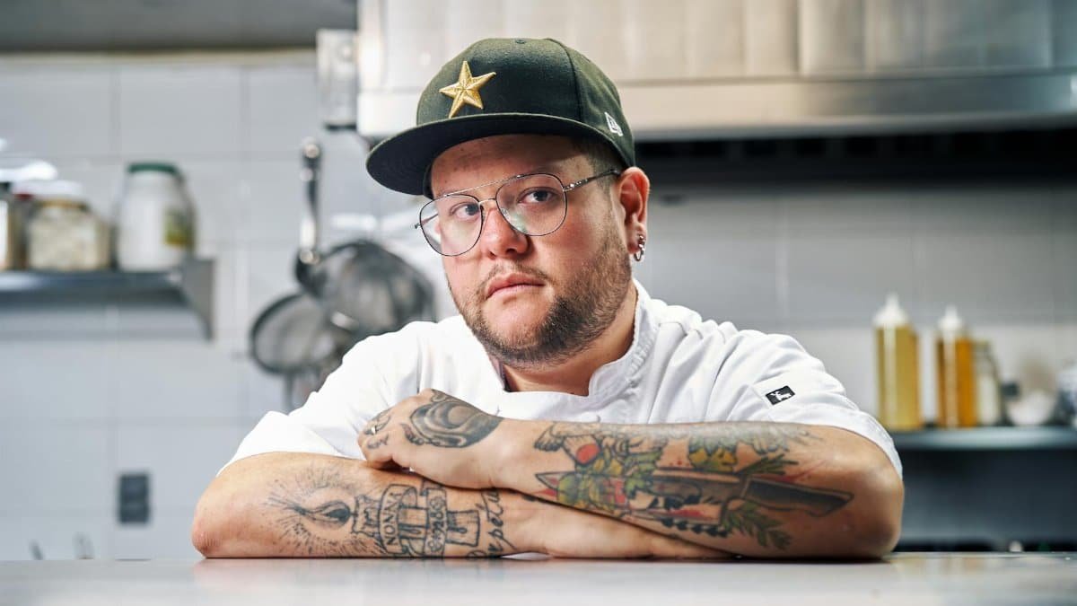 A tattooed chef poses confidently at his workstation in a professional kitchen.