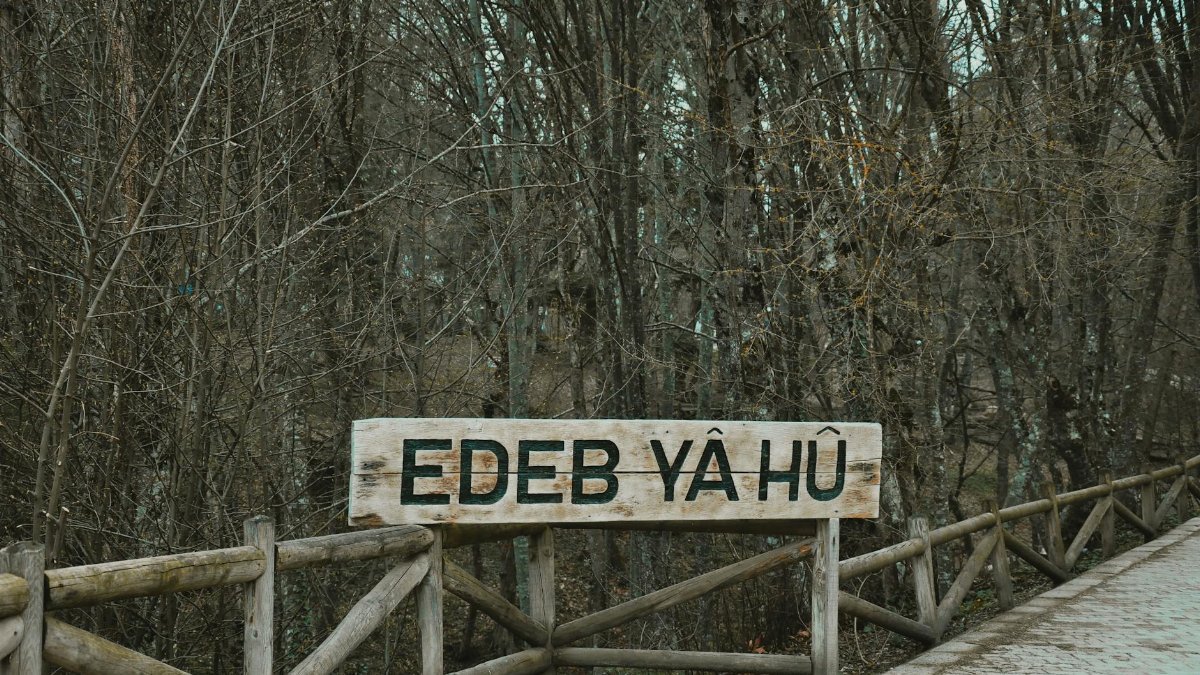 A rustic wooden sign with the text 'Edeb Yâ Hû' in a serene forest setting during fall.
