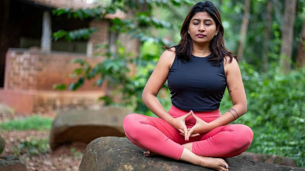 A woman practicing meditation outdoors on a rock, surrounded by lush greenery, promoting tranquility and mindfulness.
