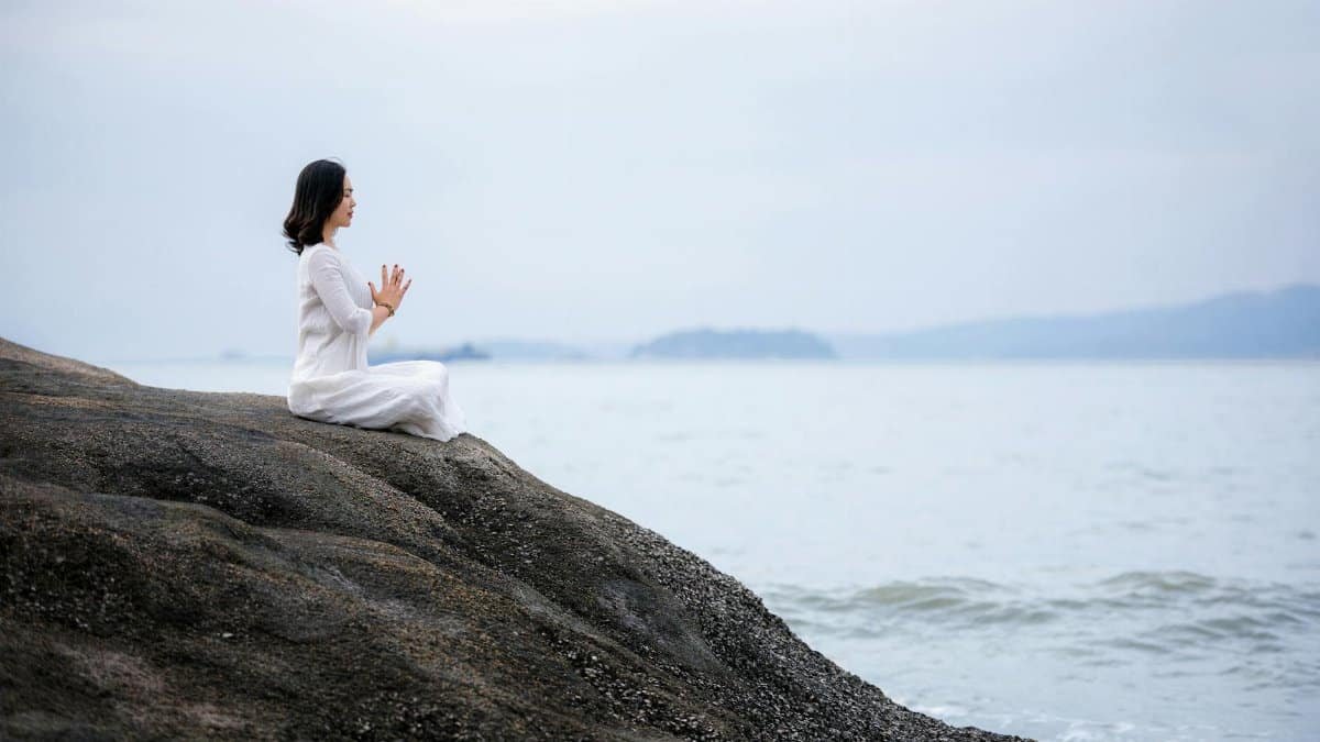 A woman meditating in a white dress by the serene sea, embodying peace and wellness.