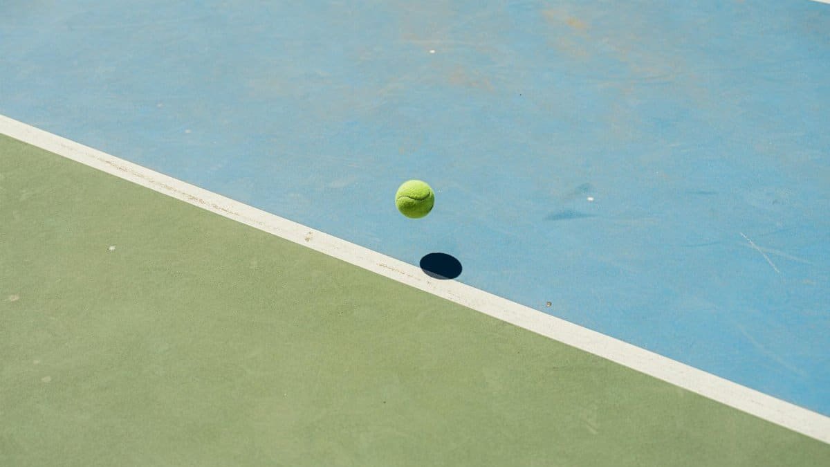A tennis ball bounces midair creating a shadow on a blue and green tennis court.