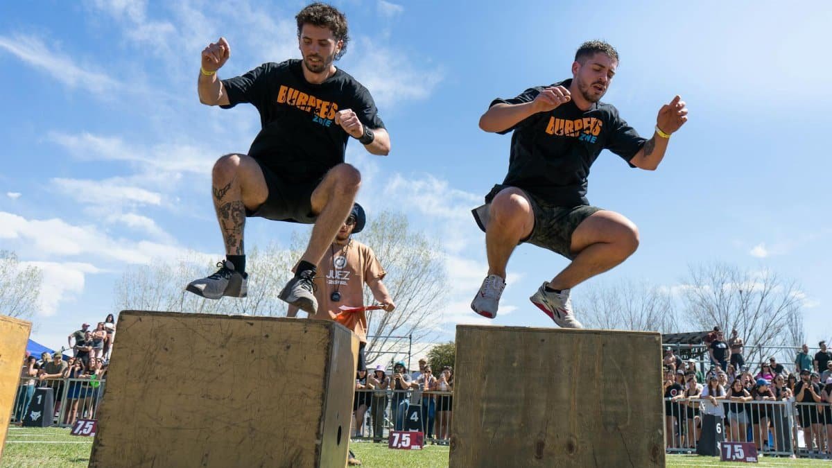Two men perform box jumps during an outdoor athletic event under a clear blue sky.